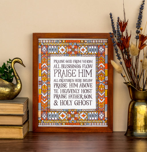 Doxology hymn art print — biblical wall art showcasing a close-up of the muted blue, red, and yellow stained glass border that surrounds the hand-lettered text, styled alongside books, vase with dried flowers and a brass swan.