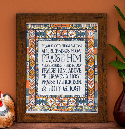 Doxology hymn art print — biblical wall art showcasing the soft blue, peach, and gold stained glass border that surrounds the hand-lettered text, framed in a dark wood frame and styled alongside a ceramic chicken and a vase of greens.