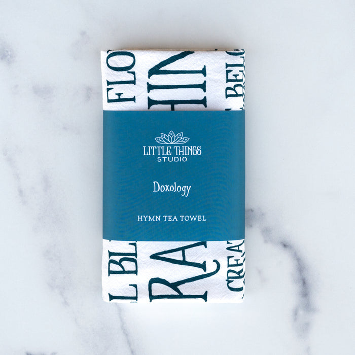 The Doxology tea towel is printed in peacock blue and surrounded by an illustrated, stained-glass inspired border, shown folded and wrapped with a belly band against a white marble background