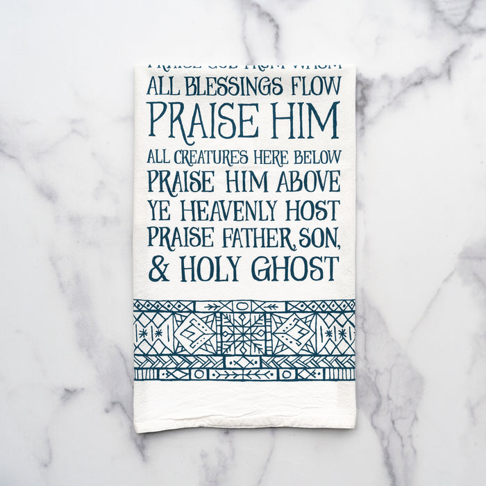 The Doxology tea towel is printed in peacock blue and surrounded by an illustrated, stained-glass inspired border, shown folded and displayed against a white marble background