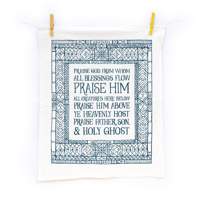 The Doxology tea towel is printed in peacock blue and surrounded by an illustrated, stained-glass inspired border, shown unfolded and pinned with clothes pins