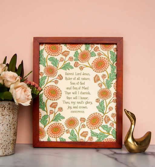 Floral detail of Fairest Lord Jesus wall art — a Christian art print showcasing hand-lettered hymn text surrounded by floral design in shades of pink, light orange and green, displayed in a dark wood frame with fresh flowers and a gold bird figure