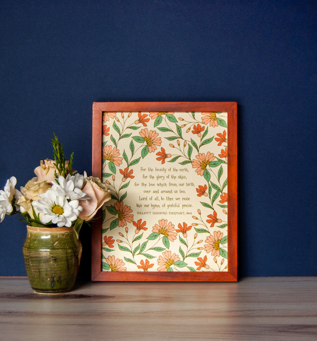 Artwork of the hymn, For the Beauty of the Earth — Christian artwork for the home that features hand-lettered hymn text surrounded by floral design in pink, yellow, light red, and green, displayed in a cherry wood frame alongside a vase of fresh flowers