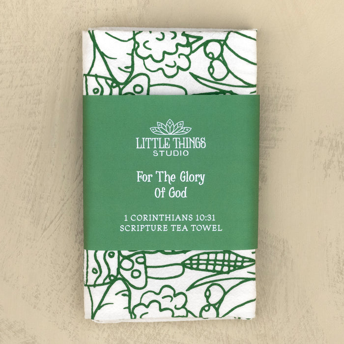 The For the Glory of God Scripture tea towel is printed in forest green and features 1 Corinthians 10:31, shown folded with a belly band for gift giving and set against a khaki background.