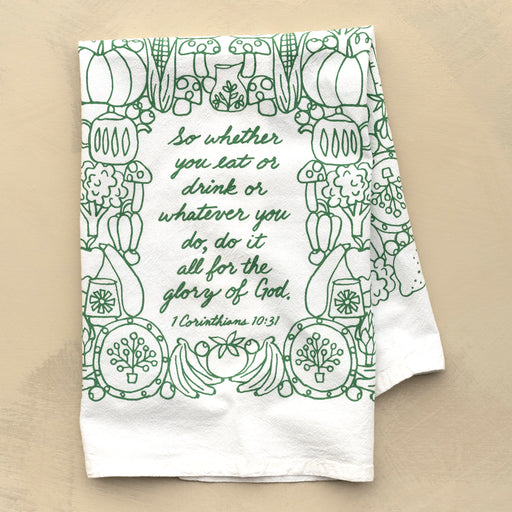 The For the Glory of God Scripture tea towel is printed in forest green and features 1 Corinthians 10:31, shown folded and set against a khaki background.