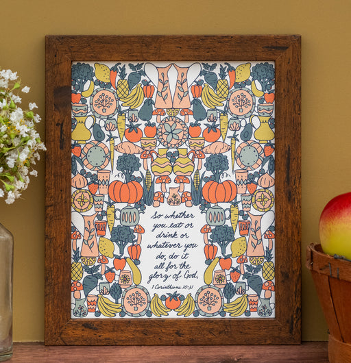 The "For the Glory of God" Scripture art print features the hand-lettered verse from I Corinthians accented by whimsical vegetable illustrations against a buttermilk background; shown in a dark wood frame and styled with a vase of flowers and a basket of fruit.