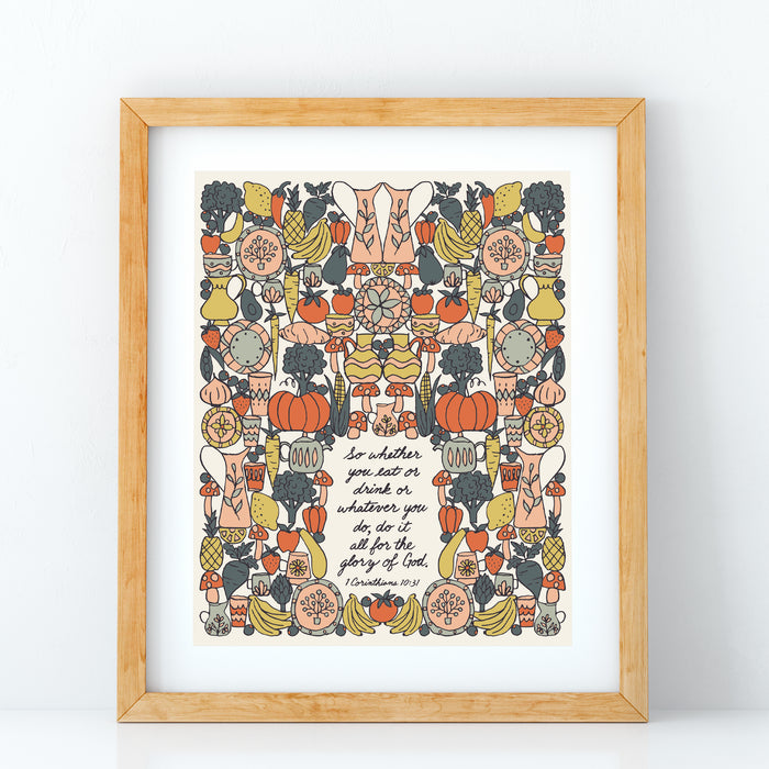 The "For the Glory of God" Scripture art print features a hand-lettered verse from I Corithians accented by whimsical vegetable illustrations against a buttermilk background; shown in a light wood frame against a white background.