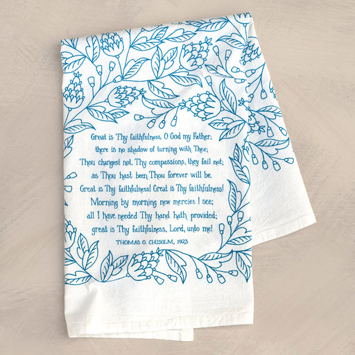 Great Is Thy Faithfulness tea towel is printed in a striking cerulean blue. The hand lettered text is surrounded by illustrated floral design, and the towel is shown folded against a khaki colored background.