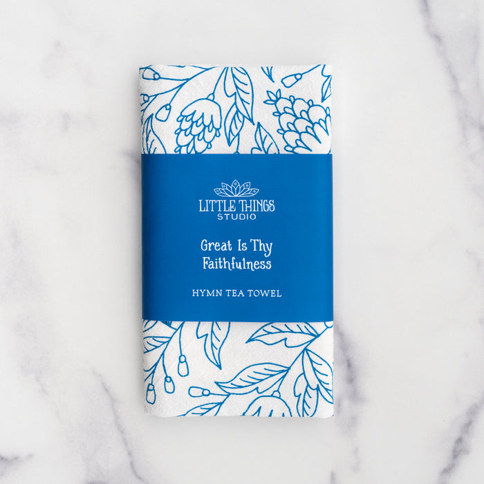 Great Is Thy Faithfulness tea towel is printed in a striking cerulean blue. The hand lettered text is surrounded by illustrated floral design, and the towel is shown folded and wrapped with a paper belly band.