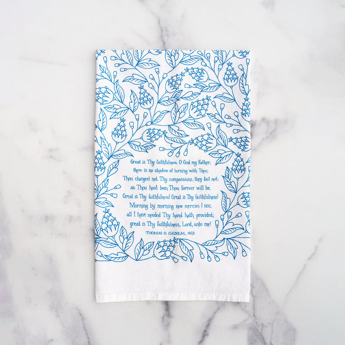 Great Is Thy Faithfulness tea towel is printed in a striking cerulean blue. The hand lettered text is surrounded by illustrated floral design, and the towel is shown folded against a white marble background.