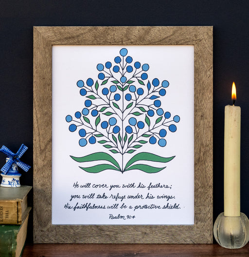 "He Will Cover You" Scripture art print features the hand-lettered verse from Psalm 91 accented by a beautiful blue and green floral against a fleecy white background. Shown here in a wooden frame, styled with a candle, books and a ceramic figure.