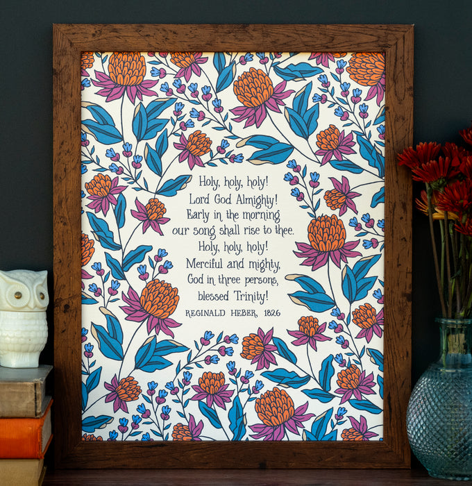 Holy Holy Holy art print — biblical wall art featuring hand lettered text surrounded by floral illustrations in shades of magenta, teal, and burnt orange, shown styled in a dark wood frame alongside an owl figurine, stack of books, and a vase of flowers.