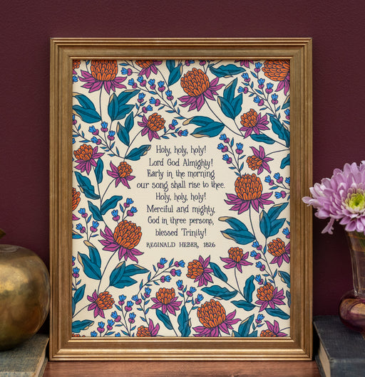 Holy Holy Holy art print — biblical wall art featuring hand lettered text surrounded by floral illustrations in shades of magenta, teal, and burnt orange displayed in a frame with vase of flowers and a gold apple.