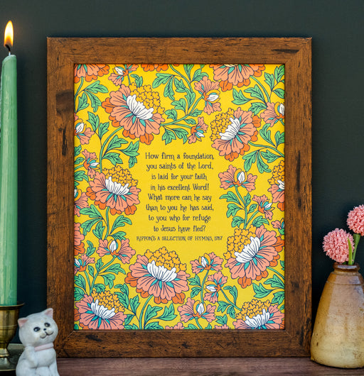 How Firm a Foundation art print — hand-lettered hymn text on a mustard background surrounded by a floral design in orange, pink, and goldenrod against a mango background, pictured in a dark wood frame and styled with a ceramic vase with a flower, candle, and ceramic animal.