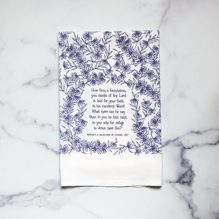 How Firm a Foundation hymn tea towel is printed in periwinkle and shown folded with a white marble background