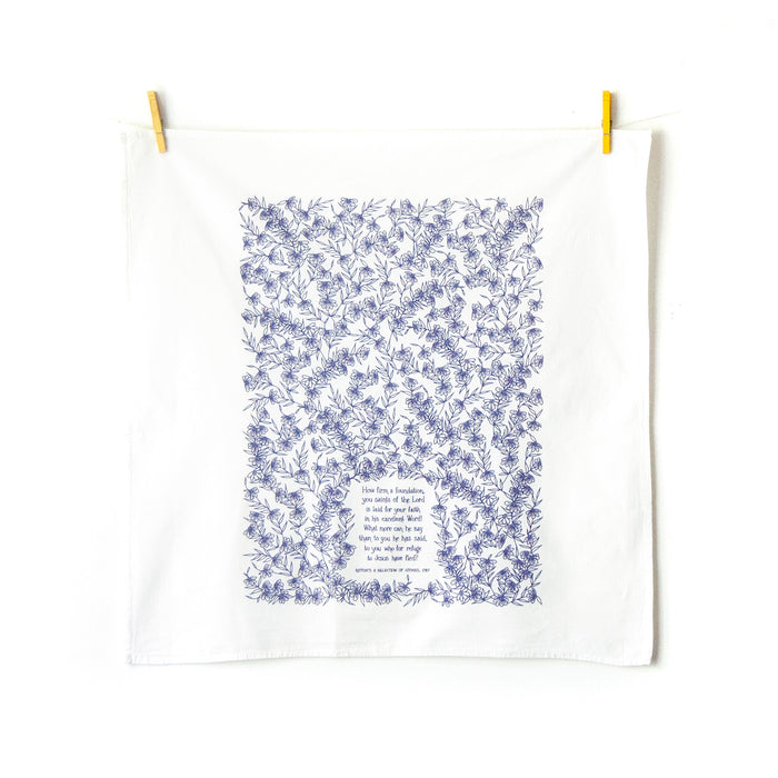 How Firm a Foundation hymn tea towel is printed in periwinkle and displayed unfolded, hanging with clothes pins