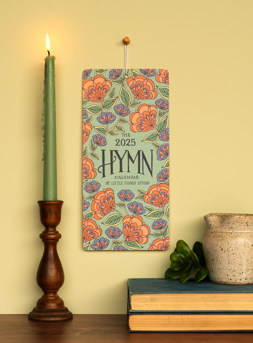 Celebrate 2025 with hymns both ancient and modern, bold floral patterns, and intricate hand-lettering, shown styled with a candle, ceramic jug, greens, and books, hung on an oatmeal toned wall.