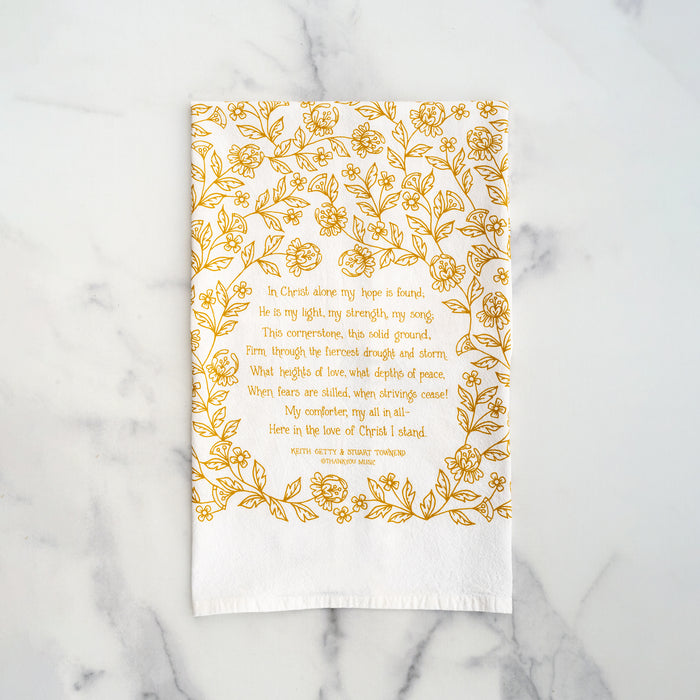 The In Christ Alone tea towel is printed in honey gold and features floral illustrations surrounding the hand-lettered hymn text. Pictured folded against a white marble background.