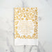 The In Christ Alone tea towel is printed in honey gold and features floral illustrations surrounding the hand-lettered hymn text. Pictured folded against a white marble background.