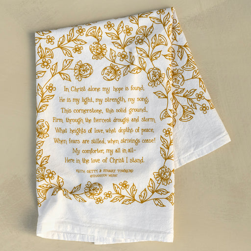 The In Christ Alone tea towel is printed in honey gold and features floral illustrations surrounding the hand-lettered hymn text. Pictured folded against a khaki background.