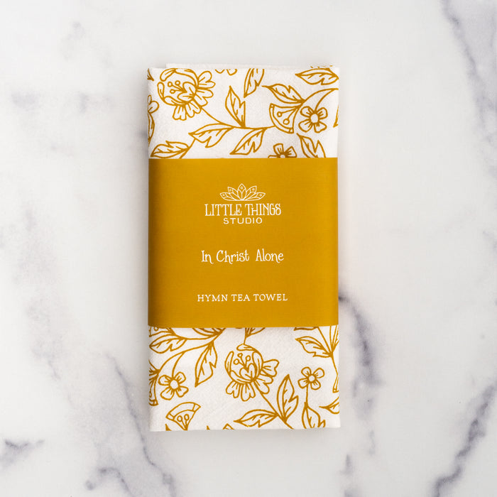 The In Christ Alone tea towel is printed in honey gold and features floral illustrations surrounding the hand-lettered hymn text. Pictured folded and wrapped with a paper belly band.