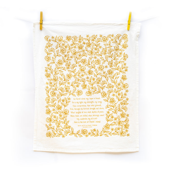 The In Christ Alone tea towel is printed in honey gold and features floral illustrations surrounding the hand-lettered hymn text. Pictured unfolded and hanging with clothes pins against a white background.