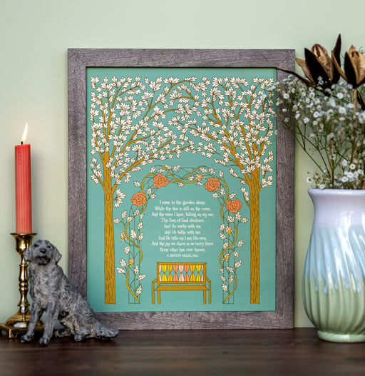 In the Garden gospel song art print features the hand lettered song text surrounded by illustration of a beautiful garden setting against a teal background, displayed in a gray wood frame, styled with a vase of flowers and greens, a candle, and a dog figurine.