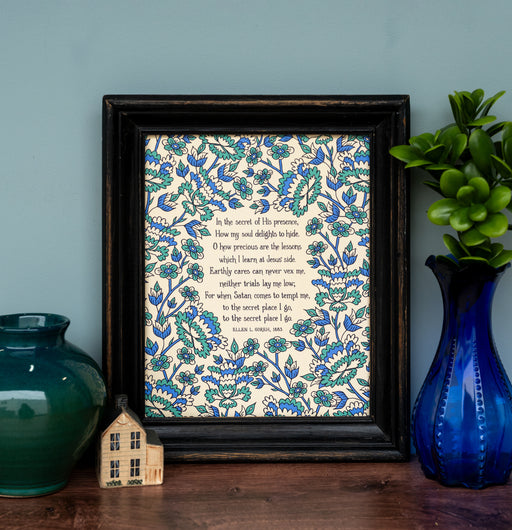 In the Secret of His Presence hymn art print in 8x10 size features hand-lettered hymn text surrounded by a vibrant blue and aqua floral illustration and shown styled with a frame, vase of greens, glass vase and wooden house figurine.