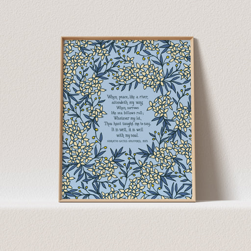 The "It Is Well" art print features the beloved hymn text surrounded by delicate floral and a sky blue background that will brighten up any room. Shown here in a light wood frame against a white background.