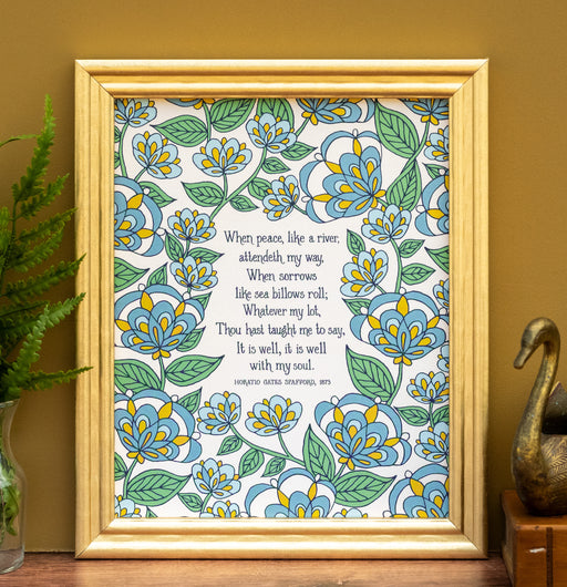 The "It Is Well" hymn art print features hymn text surrounded by a blue and green florals against a vanilla background, shown framed and styled with a vase of greens, a book, and a bronze figurine.