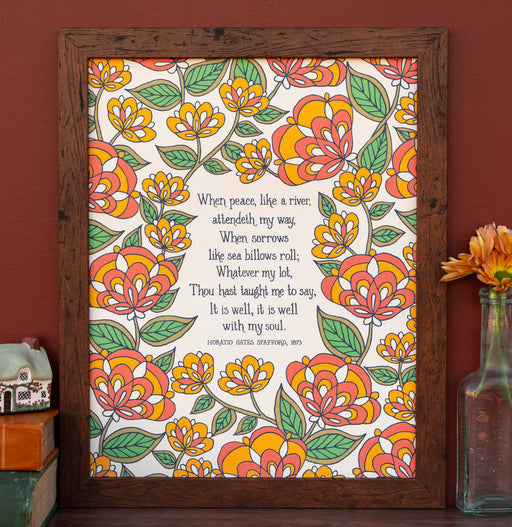 The "It Is Well" 11"x14" hymn art print features hymn text surrounded by a orange florals against a cream background, shown here in a dark wood frame alongside a vase of flowers, books, and a figurine.