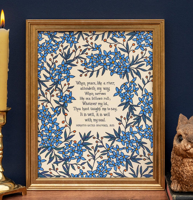 "It Is Well With My Soul" hymn art print features the beautiful hymn, surrounded by a gorgeous periwinkle florals with a creamy vanilla background; shown framed and styled with a candle and a ceramic figurine.