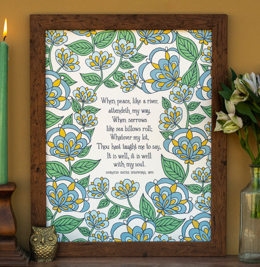 The "It Is Well" hymn art print in 11x14 features hymn text surrounded by a blue and green floral against a vanilla background; shown in a wooden frame and styled with a candle, ceramic owl, and a vase of flowers.