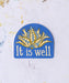 The "It Is Well" hymn sticker features an arched window shape, deep periwinkle background and floral motif in shades of blue, gold, and cream, shown against a worktable background.