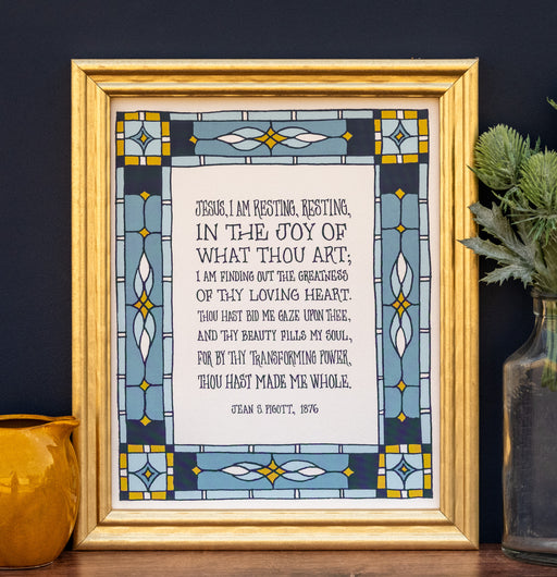 The Jesus, I Am Resting art print features a verse of the beloved hymn framed by a colorful geometric border. Shown in a gold frame and stuled with a vase of greens and a figurine.