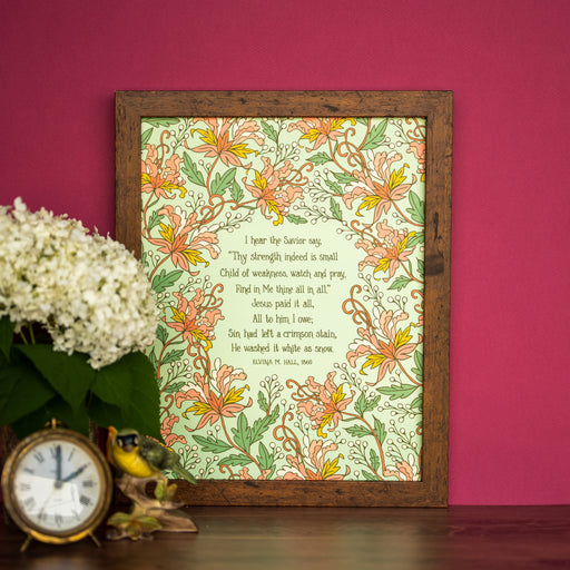 The "Jesus Paid It All" 11x14 wall art features a verse of the hymn surrounded by bold pink floral on a green background, shown here in a wooden frame and styled with a vase of flowers and a clock.