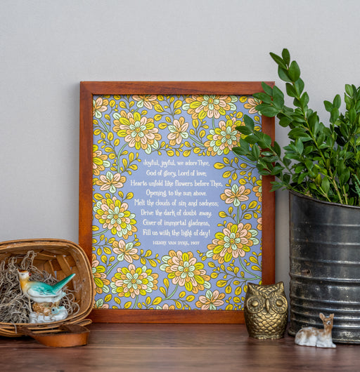 Joyful, Joyful, We Adore Thee hymn art print features bold multi-colored floral against a blue lavender background, displayed in a dark wood frame, styled alongside a tin of greens and ceramic figurines