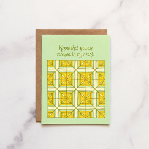 Carried in My Heart Greeting Card features he hand illustrated patchwork pattern highlighted in greens and yellows, pictured against a white marble background.