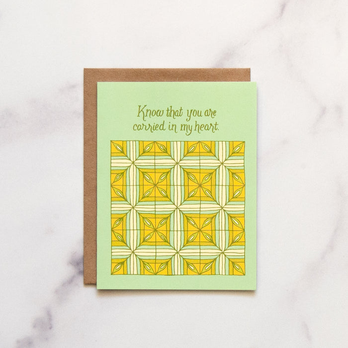 Carried in My Heart Greeting Card features he hand illustrated patchwork pattern highlighted in greens and yellows, pictured against a white marble background.