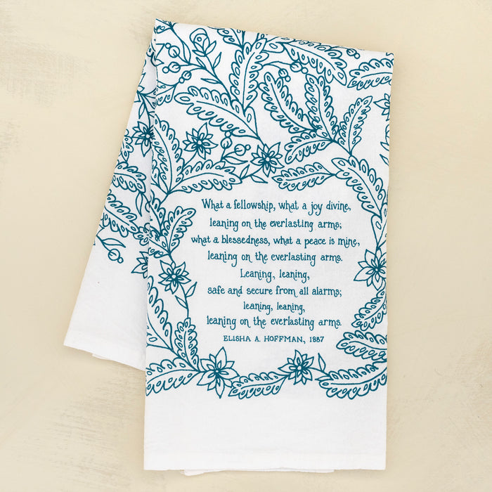 Leaning on the Everlasting Arms tea towel is printed in deep sea blue and features hand lettered hymn text surrounded by illustrated florals, pictured here folded against a khaki background.