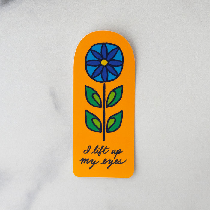 The “Lift My Eyes” Scripture sticker features hand-lettered text from Psalm 121:1 and reads "I lift up my eyes." The words are surrounded by a vibrant floral against a rich sunset orange background, shown here against a gray marble background.