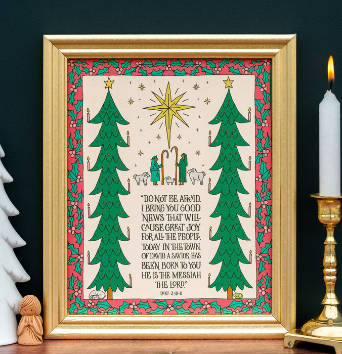 The "Good News of Great Joy" art print, which features the hand-lettered text of Luke 2:10–11 surrounded by festive trees and the wondering shepherds, is shown in a gold frame and styled with a candle and white ceramic tree and angel carving.