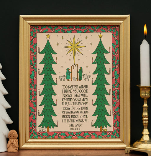 The "Good News of Great Joy" art print, which features the hand-lettered text of Luke 2:10–11 surrounded by festive trees and the wondering shepherds, is shown in a gold frame and styled with a candle and white ceramic tree and angel carving.
