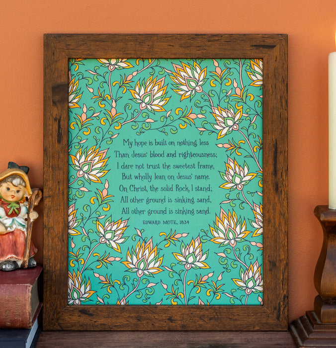 The "My Hope Is Built" hymn art print features a verse from the hymn surrounded by a delicate multicolored floral illustration against an aquamarine background, will compliment any room; shown here framed and styled with books, a ceramic figure, and a candle.