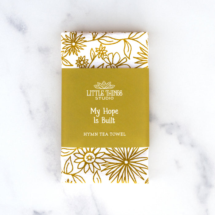 The Hope is Built on Nothing Less tea towel is printed in Sicilian green and features the hand lettered hymn text surrounded by intricate floral illustrations, pictured folded and wrapped with a paper belly band for gift giving