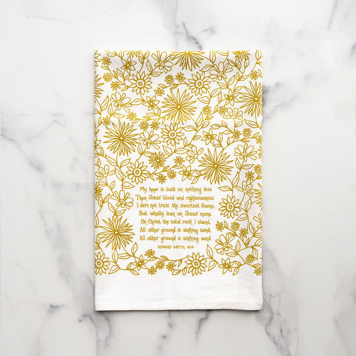 The Hope is Built on Nothing Less tea towel is printed in Sicilian green and features the hand lettered hymn text surrounded by intricate floral illustrations, pictured folded against a white marble background