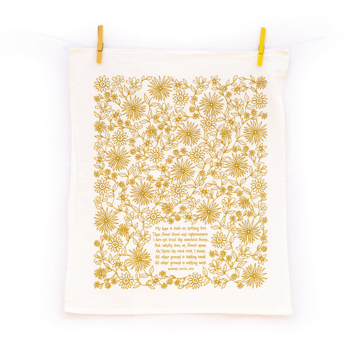 The Hope is Built on Nothing Less tea towel is printed in Sicilian green and features the hand lettered hymn text surrounded by intricate floral illustrations, pictured unfolded and hanging with clothes pins