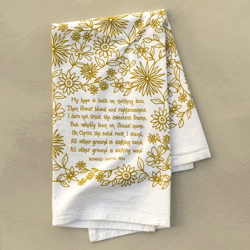 The Hope is Built on Nothing Less tea towel is printed in Sicilian green and features the hand lettered hymn text surrounded by intricate floral illustrations, pictured folded against a khaki background