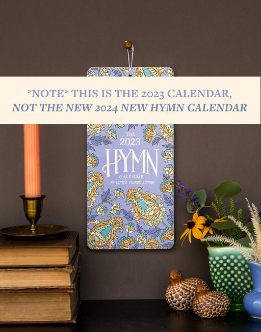 2023 Hymn Calender, shown styled with books, candle in holder, vase with greens and figurines, as well as a banner clarifying that this is NOT the new 2024 Hymn Calendar.