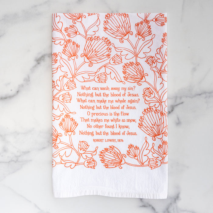 The Nothing But the Blood hymn tea towel is printed in sunset orange and features lyrics from the hymn accented by intricate floral illustration, shown folded against a gray marble background